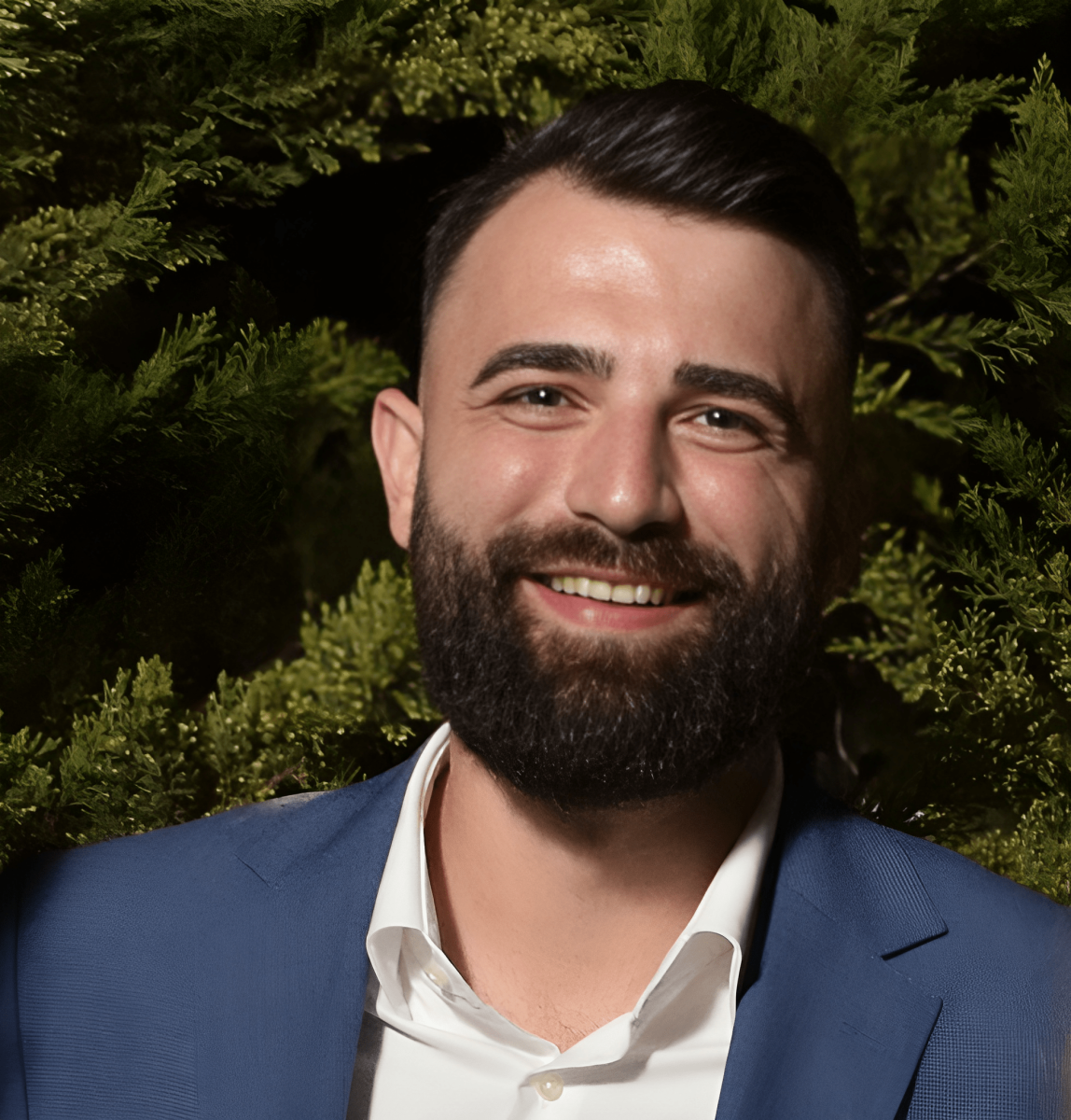 Image of the author, Adrian Nita. A head and shoulder shot of a fair skinned man with brown hair, a brown beard and a light blue suit with a white shirt, against a green bushy background.