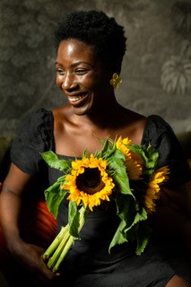 Portrait of Marie Kacouchia. A Black woman with short hair, wearing a black puffed short sleeved dress. Marie is holding sunflowers close to her chest.