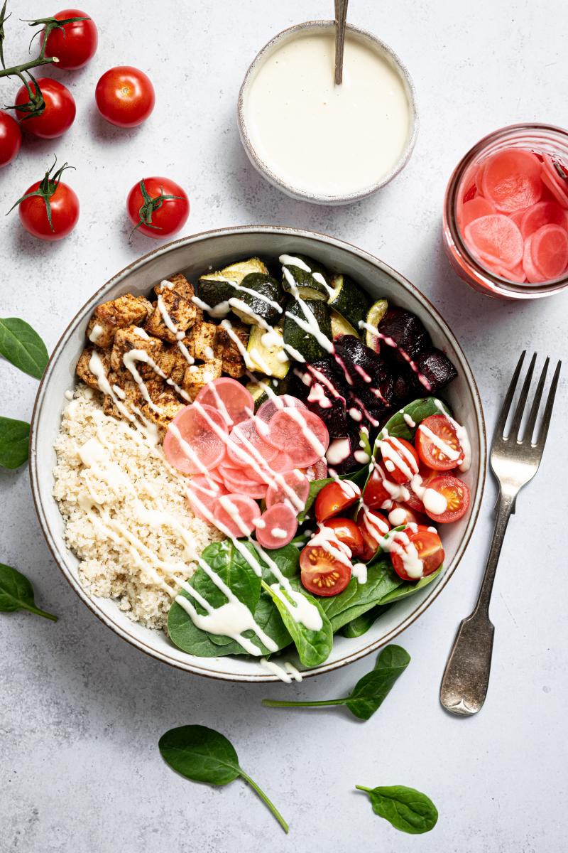 Separated into portions in a bowl; fonio, courgette, radish, tofu, pickles, tomatoes and spinach. The bowl is surrounded by the ingredients in the bowl.